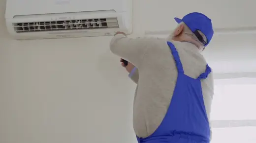 An elderly handyman in blue overalls repairs a white wall-mounted air conditioner.