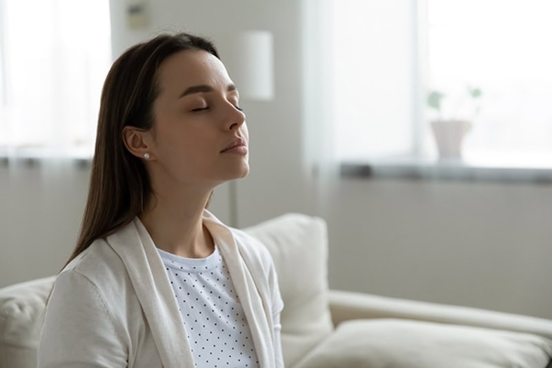 Why Do I Need a Humidifier? Woman sitting on couch with eyes closed.|Why Do I Need a Humidifier? Woman sitting on couch with eyes closed.