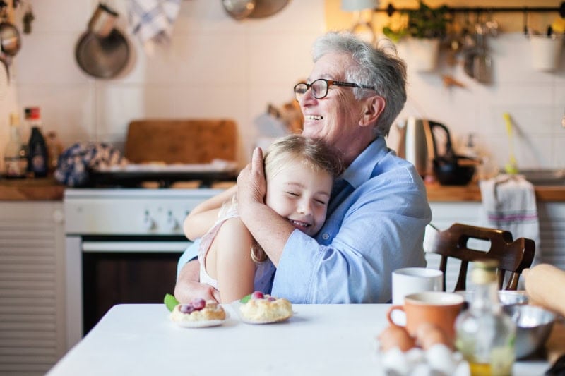 Happy grandmother is hugging granddaughter in cozy home kitchen. Family is cooking together. Senior woman and cute little child girl are smiling. Kid is enjoying kindness