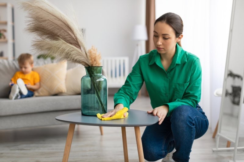 How to Reduce Dust in Your Home. Young mom dusting off coffee table and tidying up in living room while her toddler son watching cartoon on cellphone
